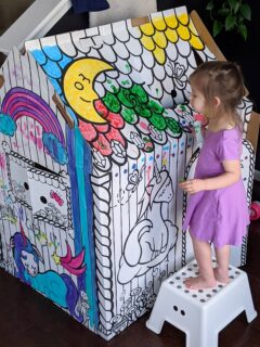 little girl coloring on and painting a cardboard colorable playhouse