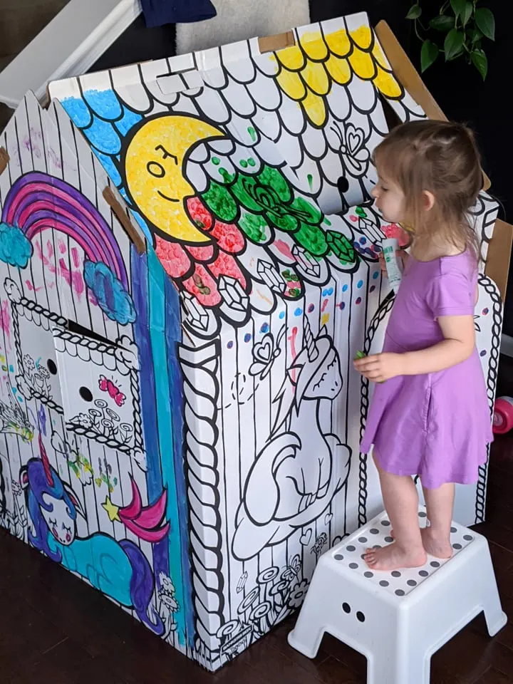 little girl coloring on and painting a cardboard colorable playhouse