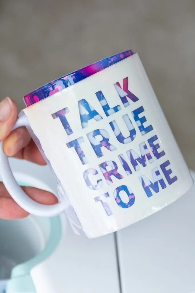 removing an Infusible Ink transfer sheet from a mug that says "talk true crime to me" created with a Cricut Mug Press