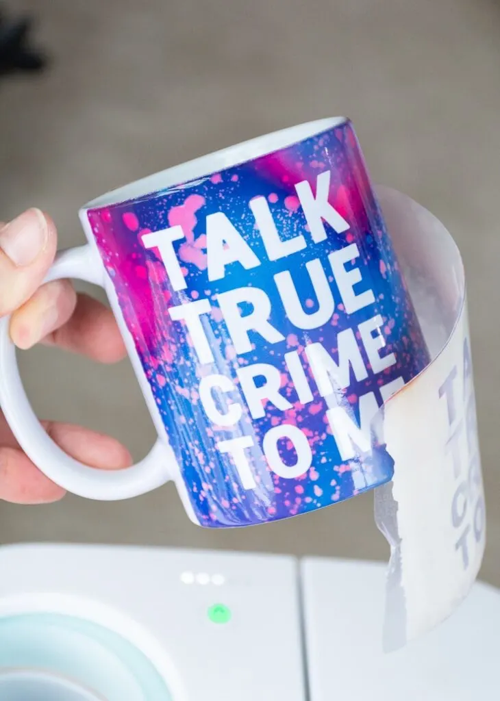 removing an Infusible Ink transfer sheet from a mug that says "talk true crime to me" created with a Cricut Mug Press